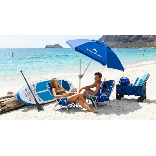 A smiling couple relaxing in blue Tommy Bahama beach chairs on a Maui beach, surrounded by an inflatable stand-up paddleboard, snorkel gear, boogie boards, a beach umbrella, and a loaded cargo wagon.